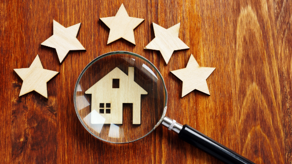 Ein Holzhaus wird durch eine Lupe vergrößert, umgeben von fünf Holzsternen auf einer hölzernen Oberfläche.